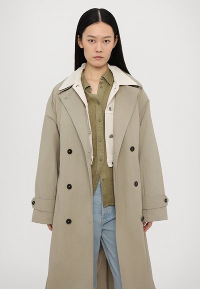 Beige trench coat with large collar and double-breasted front, paired with a light green button-up shirt. Model stands against a white background.