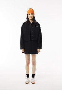 Veste noire courte avec de grands boutons et des poches latérales, associée à une jupe noire. Bonnet orange à côtes, baskets blanches avec des accents marron.