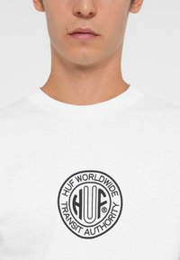 Balta kokvilnas T-kreklu, kas dekorēts ar apaļu melnu logotipu ar tekstu "HUF WORLDWIDE TRANSIT AUTHORITY" un atšķirīgu fontu centrā.