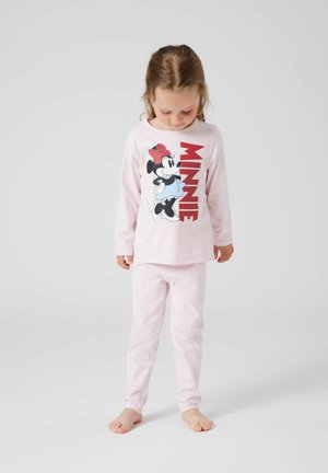 Jeune fille portant un pyjama Minnie Mouse rose clair, debout pieds nus sur un fond blanc uni, regardant vers le bas.