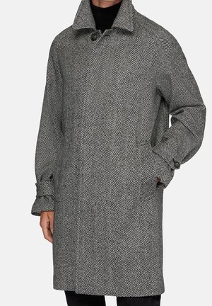 Homme portant un pardessus à motif chevrons noir et blanc, col relevé et mains partiellement dans les poches.