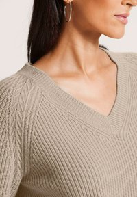 Frau, die einen beigefarbenen gestrickten V-Ausschnitt-Pullover mit Zopfmuster-Details an den Schultern und silbernen Creolen trägt.