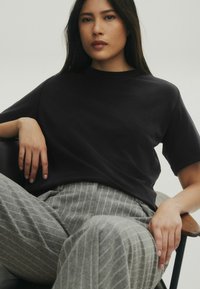 Schwarzes T-Shirt mit kurzen Ärmeln und entspannter Passform, kombiniert mit grauen gestreiften Hosen. Glatte Textur, minimalistisches Design, sitzende Pose auf einem Stuhl.