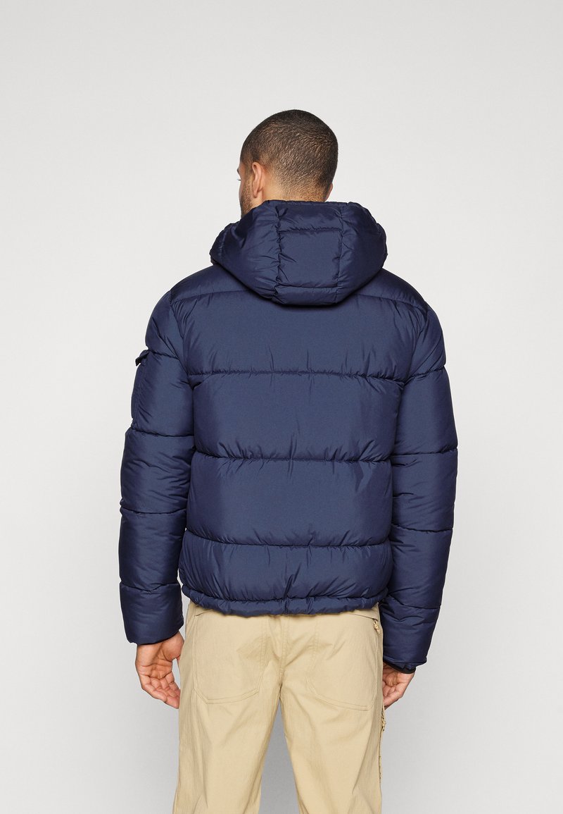 Mann in einer marineblauen Steppjacke mit Kapuze, von hinten stehend, kombiniert mit beigen Hosen vor einem schlichten Hintergrund.