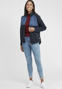 Veste zippée marine avec panneau matelassé bleu sur la poitrine, en tissu doux, dotée de deux poches latérales, portée sur un haut bordeaux, associée à un jean bleu clair et des chaussures blanches.
