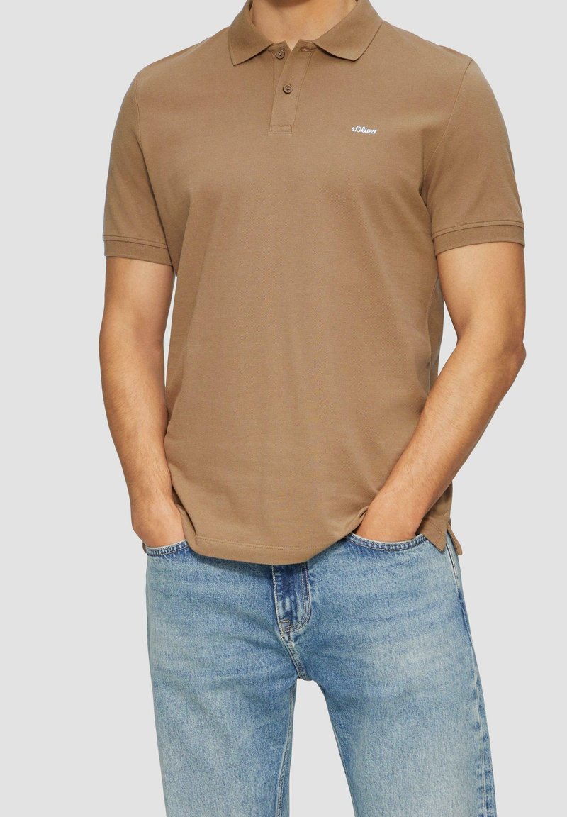 Polo shirt marron en coton, avec un boutonnière, des manches courtes et un logo sur le côté gauche. Porté avec un jean bleu clair.