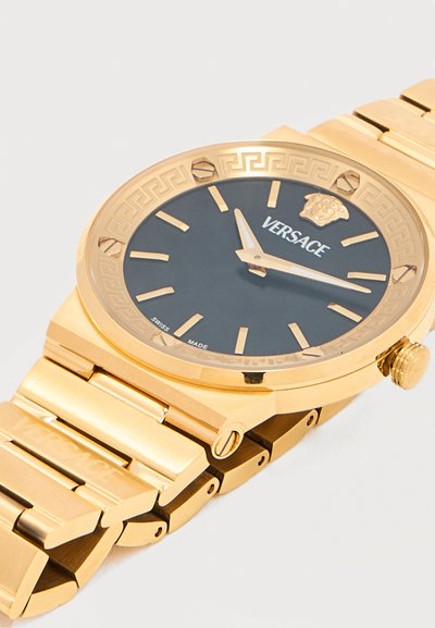 Ρολόι χειρός Versace σε χρυσαφί απόχρωση με μαύρο καντράν, χρυσαφί δείκτες ώρας, λογότυπο Versace με κεφάλι και μπρασελέ από συνδεδεμένα μεταλλικά στοιχεία.