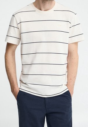 Uomo che indossa una maglietta crema a maniche corte con sottili strisce orizzontali nere e pantaloni blu scuro con le mani nelle tasche.