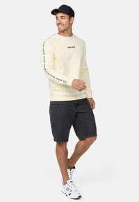 Sudadera de manga larga color crema con logo negro, combinada con shorts de mezclilla oscuros. Los accesorios incluyen una gorra negra y zapatillas blancas.