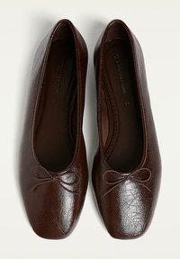 Ballerines en cuir marron avec une surface texturée, un bout rond et un petit nœud décoratif sur le devant. L'intérieur présente un motif à pois.