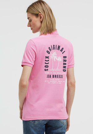 Frau, seitlich stehend, trägt ein pinkes Poloshirt mit der Aufschrift „SOCCX Original Brand Sea Breeze“ und Palmen-Grafiken auf dem Rücken.
