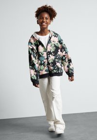 Sudadera con cremallera de flores en negro con patrones en rosa y verde, que cuenta con un bolsillo tipo canguro y puños acanalados. Combinada con pantalones anchos blancos.