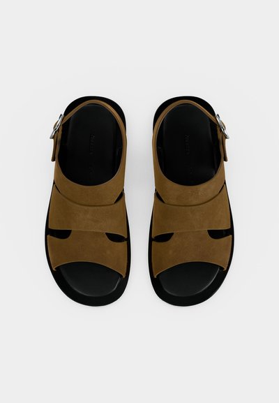 Ζευγάρι καφέ δερμάτινα σανδάλια με μαύρες σόλες, με φαρδιές λουριά και μεταλλικά κούμπωμα, από ψηλά σε λευκό φόντο.