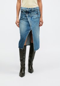 Η τζιν φούστα διαθέτει μοναδικό σχέδιο τύλιξης, ανοιχτό μπλε χρώμα, μεσαίο μήκος με άνοιγμα στο μπροστινό μέρος και αντίθετες τζιν τσέπες.