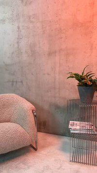 Fauteuil boucle beige incurvé à côté d'une table d'appoint métallique en fil supportant une plante en pot et des magazines empilés contre un mur en béton.