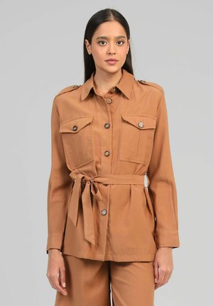 Jeune femme portant une veste-chemise marron boutonnée avec des poches à rabat et une ceinture nouée à la taille, associée à un pantalon marron assorti.