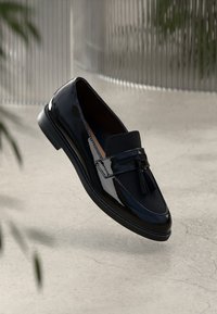 Mocassino in pelle verniciata nera con un accento a nappina, caratterizzato da una superficie liscia, punta arrotondata e design elegante su uno sfondo chiaro e tessurizzato.