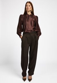 Bruine satijnen blouse met een grote kraag en een gestrikt detail, gecombineerd met hooggetailleerde bruine plooibroek en spitse bruine hakken.