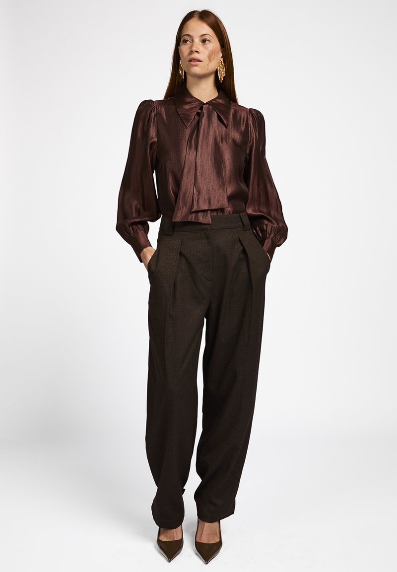 Bruine satijnen blouse met een grote kraag en een gestrikt detail, gecombineerd met hooggetailleerde bruine plooibroek en spitse bruine hakken.