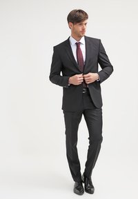 Svart skreddersydd dress med en enkelt knappjakke, parret med en hvit dresskjorte og et mørkerødt prikket slips, fullført med svarte sko.