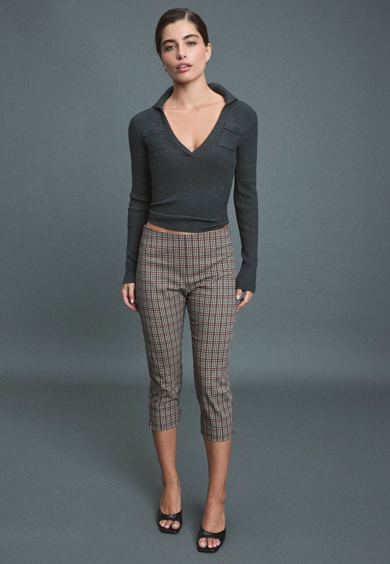 Femme debout portant un haut à manches longues gris foncé avec col en V, un pantalon court à carreaux et des talons noirs ouverts à l'avant, sur un fond gris uni.