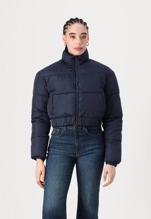 Veste sans manches matelassée bleu marine avec un col haut et une fermeture éclair. Comprend des poignets élastiques et une silhouette ample. Portée avec un jean en denim taille haute.