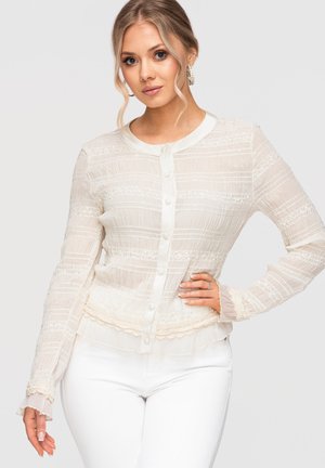 Kobieta o blond włosach nosi przezroczystą białą bluzkę z długim rękawem, guzikami, koronkowymi detalami i białe spodnie, pozując z jedną ręką na biodrze.