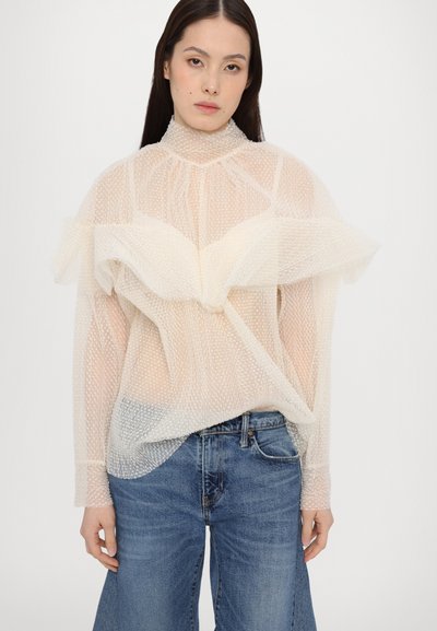 Διαφανής, υφασματοποιημένη λευκή μπλούζα με ψηλό λαιμό και φάσες, συνδυασμένη με μπλε τζιν παντελόνι σε φαρδιά γραμμή.