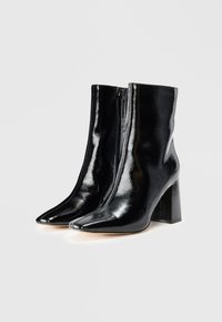 Botas de tobillo de charol negro con punteras afiladas, cremallera lateral y tacones cuadrados, que presentan un acabado brillante y elegante.