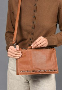 Bandolera de cuero marrón con cierre de cremallera, adornada con tachuelas metálicas en la parte delantera. Forma plana y rectangular con una correa ajustable.