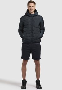Donkerblauwe pufferjack met capuchon, met een gestikte ontwerp, ritsborstzak en zwarte accenten. Draagt over zwarte shorts en laarzen.