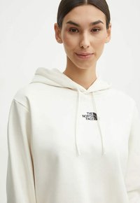 Biała bluza z kapturem wykonana z miękkiego materiału, z przednią kieszenią i regulowanym kapturem. Na lewej piersi znajduje się czarne logo The North Face.