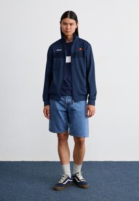 Marineblå zip-up jakke med høy krage, med hvite og røde detaljer. Brukt over en marineblå T-skjorte og kombinert med denimshorts og marineblå sneakers.