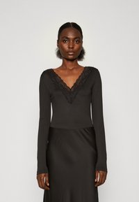 Must have pikkade varrukatega must bodysuit, millel on sügav V-kujuline kaelus koos pitsäärisega. Pehme ja sile materjal, mis on vormitud disaini ja elegantse siluettiga.