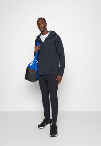Felpa con zip blu navy e pantaloni abbinati, borsa da palestra nera e blu, e scarpe sportive nere. Materiale liscio con vestibilità comoda.