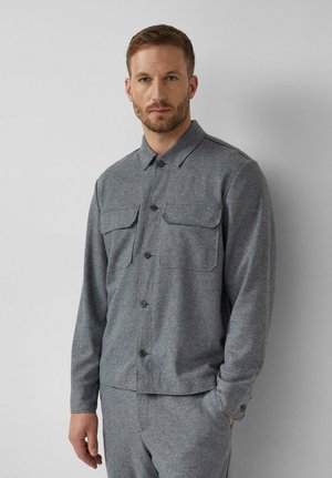 Mann trägt ein graues Hemd mit Knöpfen und zwei Brusttaschen sowie passende graue Hosen, steht vor einem einfarbigen hellen Hintergrund.