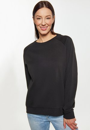 Sweatshirt noir à manches longues avec une texture côtelée, col rond et manches raglan, associé à un jean en denim bleu clair.