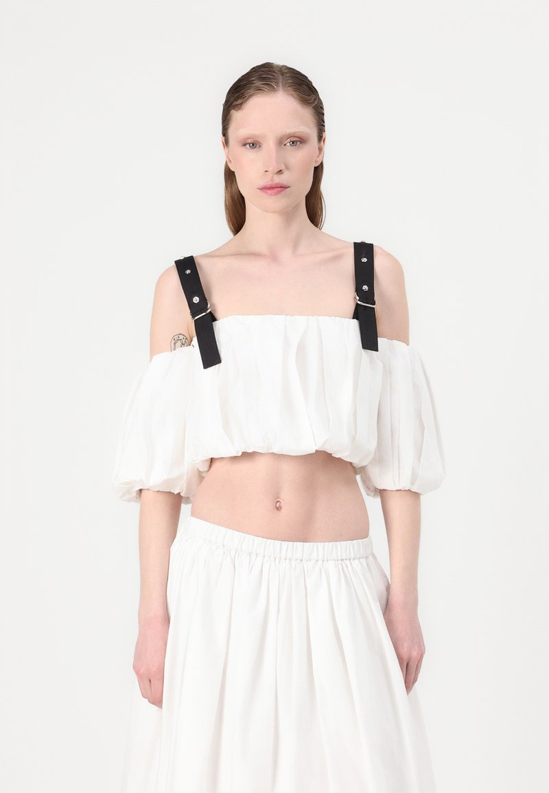 Blusa bianca corta con maniche a sbuffo, dotata di cinturini in pelle nera e accessori in argento. Abbinata a una gonna bianca fluida coordinata.