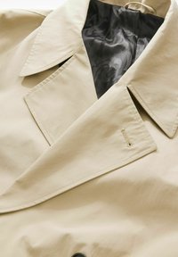 Manteau beige avec un large col, un devant double boutonnage, des coutures visibles et une doublure intérieure lisse avec un motif subtil.