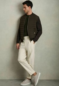 Jachetă bomber cu un corp maro matlasat și mâneci întunecate, combinată cu pantaloni plisati crem și adidași gri deschis, stând în fața unui fundal estompat.