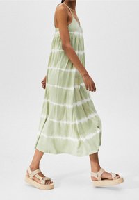 Robe maxi sans manches vert clair avec des rayures blanches effet tie-dye, coupe large et col en V. Associée à des sandales à semelle compensée beige avec des lanières.