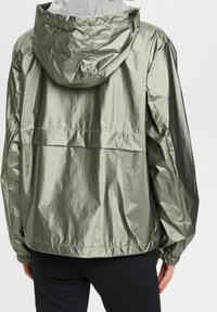 Zielona, lekka kurtka przeciwwiatrowa z kapturem, charakteryzująca się błyszcząca, pomarszczoną fakturą oraz elastycznymi mankietami, plisą z tyłu i ściągaczem w pasie.