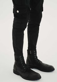 Črni usnjeni bojevski škornji z gladkim teksturo, zavezovanje z vezalkami ter detajlom z zadrgo. Kombinirani z oprijetimi črnimi cargo hlačami s stranskimi žepi.