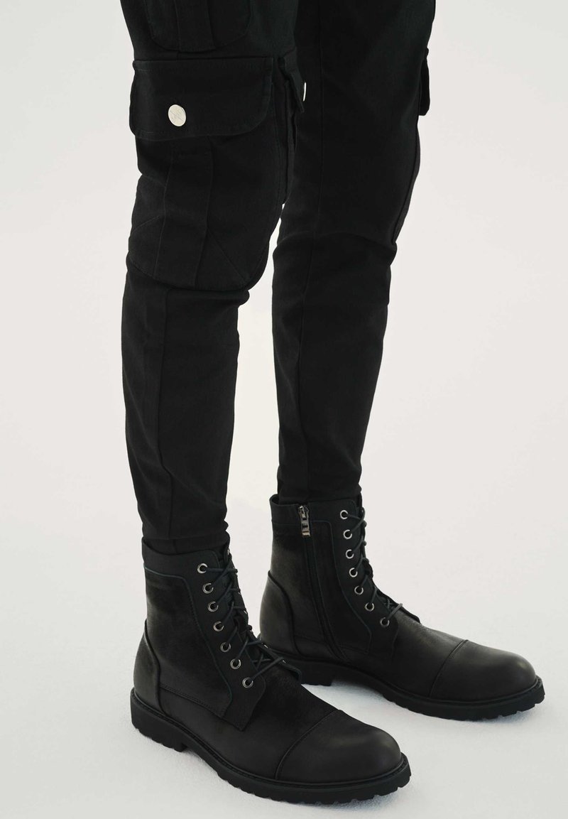 Črni usnjeni bojevski škornji z gladkim teksturo, zavezovanje z vezalkami ter detajlom z zadrgo. Kombinirani z oprijetimi črnimi cargo hlačami s stranskimi žepi.