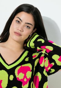 Schwarzer Pullover mit V-Ausschnitt, der ein auffälliges Muster aus pinken Totenköpfen und neon grünen Akzenten aufweist. Hergestellt aus strukturiertem Strickstoff, mit gestreiften Bündchen.