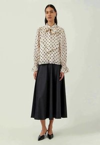 Beige Bluse mit gepunktetem Muster und einer Schleife am Hals, kombiniert mit einem schwarzen satinlangen Rock. Schwarze spitze High Heels runden den Look ab.