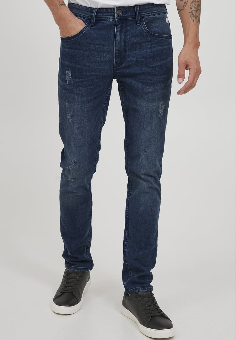 Jeans in denim blu navy con vestibilità slim, caratterizzati da toppe strappate, cinque tasche e chiusura con bottone. Abbinati a sneakers nere.