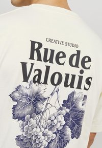 Hvit bomull T-skjorte med marineblå grafisk trykk av druer og blader. Har teksten "Rue de Valouis" iøynefallende på baksiden.