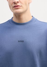 Modrá mikina vyrobená z hladkej tkaniny, ktorá má rebrovaný golier a jemné logo "BOSS" v tmavšej farbe na hrudi.