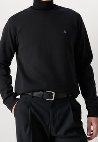 Černá mikina s dlouhým rukávem a kulatým výstřihem, s malým logem na hrudi. Látka působí hladce a jemně v kombinaci s upravenými kalhotami.